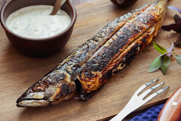 Tasty grilled mackerel on a wooden board next to a white sauce in a ceramic cup, basil sprigs, fork and kinfe, closeup