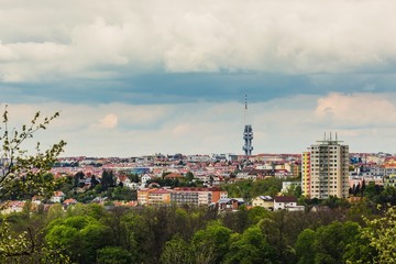 Fototapeta na wymiar Panoramic view of cityscape of spring Prague, Czech Republic, and its landmark Zizkov television tower. Grey stormy clouds sky, colorful houses and buildings, green vegetation in foreground