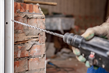 Male hands of an employee hold in their hands an electric perforator with a drill with the help of which a hole is made in the brick wall through the frame to install and fix the PVC window.