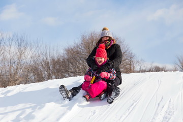 Fototapeta na wymiar Mother and little girl screaming with joy while sliding on a snow-covered hill in winter clothes during a bright sunny winter day, Beaubien Park, Montreal, Quebec, Canada