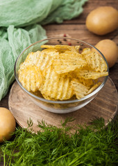 Homemade potato crisp chips inside glass bowl with fresh raw dill on wooden background and raw potatoes.