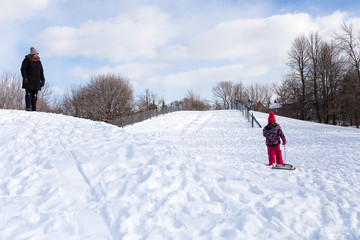 Fototapeta na wymiar Mother watching little girl with sledge climbing snow-covered hill during a bright sunny winter day, Beaubien Park, Montreal, Quebec, Canada