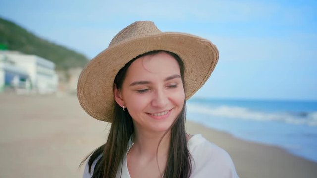 Portrait of young beautiful woman standing on beach and feeling pleasant positive emotions, smiling while sea breeze gently touch her brunette long hair, dressed in straw summer hat and white shirt