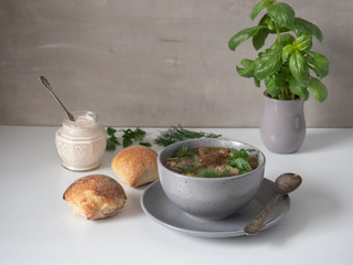 Mushroom soup in a deep bowl and dark flour bread rolls on a white table. Pot with fresh basil.
