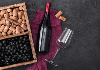 Bottle of red wine on red cloth with empty glass and dark grapes with corks and corkscrew inside vintage wooden box on dark wooden background.Top view
