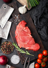 Fresh raw organic slice of braising steak fillet on chopping board with fork and knife on black stone background. Red onion, tomatoes with salt and pepper.