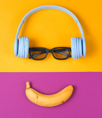 Minimalism flat lay concept. Smiling face listens to music. Headphones, 3D glasses, banana on purple yellow background. Top view