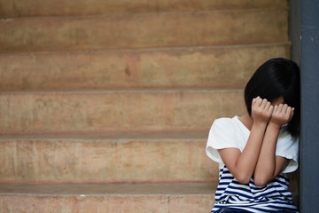 An Asian girl sat unhappy with her hands covering her face and leaning against the wall. - Powered by Adobe