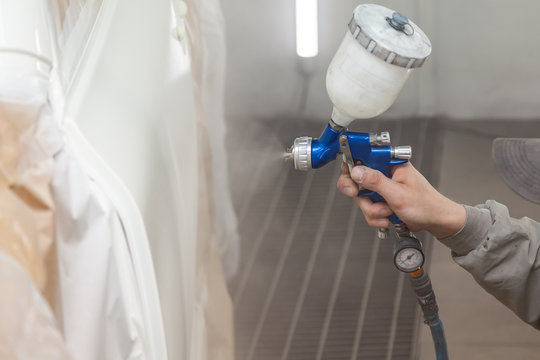 A Male Worker Paints With A Spray Gun A Part Of The Car Body In White After Being Damaged At An Accident. Door From The Vehicle During The Repair In The Workshop. Auto Service Industry Professions