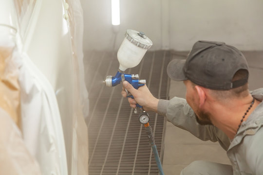 A male worker paints with a spray gun a part of the car body in white after being damaged at an accident. Door from the vehicle during the repair in the workshop. Auto service industry professions