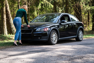 Young woman standing next to a broken car on countryside. Woman's car broken. Woman trying to fix a broken car. Despaired woman trying to fix broken car.