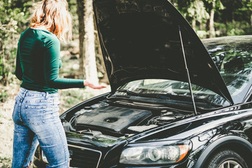 Young woman standing next to a broken car on countryside. Woman's car broken. Woman trying to fix a broken car. Despaired woman trying to fix broken car.