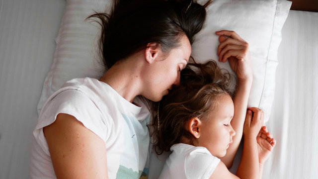 Family portrait of young mom is sleeping with her cute little daughter together on white linen in bed.