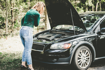 Young woman standing next to a broken car on countryside. Woman's car broken. Woman trying to fix a broken car. Despaired woman trying to fix broken car.