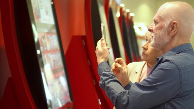 Seniors buy movie ticket from vending machine at mall. Senior man use mobile phone to read information about online. Making photos, download info from internet. Active modern life after retirement