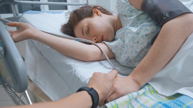 The woman lies in a hospital room, holding the railing, she convulses. The hand includes a catheter, measure the pressure, the man holding her hand. Woman preparing for surgery and childbirth. 4K