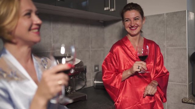 Two women chatting in the kitchen and drinking red wine from glass - One wearing blue morning gown, the other red robe dress - Laughing and smiling