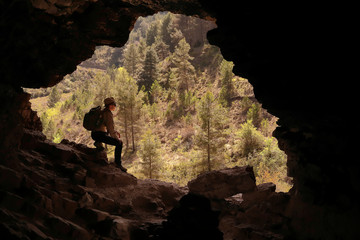 HIKER ADVENTURER MOUNTAINEER MAN  WITH HAT AND BACKPACK RESTING AT THE ENTRANCE OF A CAVE IN A VALLEY WITH MANY GREEN TREES
