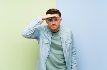 Redhead man over colorful background looking far away with hand to look something
