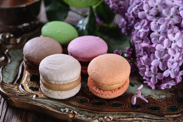 Multicolored bright macaroons on a wooden vintage tray next to flowers of lilac and a cup of tea on a wooden table, closeup