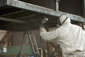 A man in a white uniform applies paint with a spray gun on a metal product.