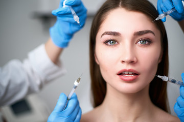 Hands of beauticians holding syringes around doll like woman face ready for injection in cosmetology clinic. Female model under influence of cosmetic treatment industry standardized beauty concept.