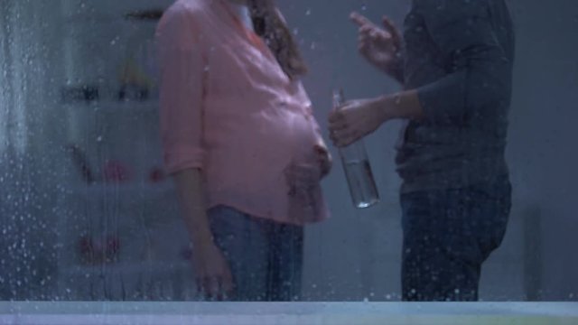 Drunk man with bottle in hands threatening pregnant woman behind rainy window