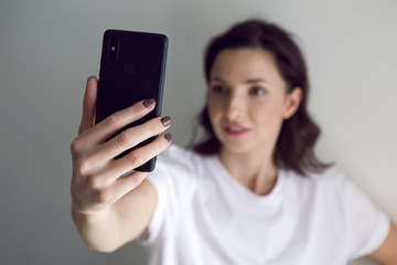 girl in a white t-shirt takes a selfie on a black phone against the white wall