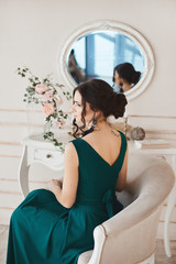 Fashionable brunette woman with stylish hairstyle in stylish evening dress and in trendy shoes sits at the dressing table