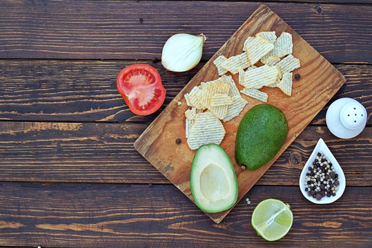 Ingredients for cooking guacamole on a wooden background: cut avocado, onion, tomato, salt, pepper, lime, chips for serving. Top view, copy space.