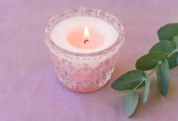Obraz na płótnie Canvas Pink burning candle in elegant crystal candle holder green silver dollar eucalyptus branch on lilac cotton table cloth. Romantic holiday table setting elements wellness interior decoration concept