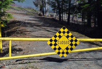Yellow and back closed sign on a gate to a campground. Blue lake and green forest in the background.