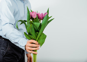 The man is holding in the hands of purple tulips on a light background. The concept of handing flowers to a woman, girl. A man wearing a shirt is holding flowers in his hands.