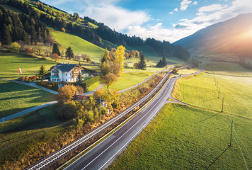 Obraz premium Aerial view of the road in mountain valley at sunset in summer in Dolomites, Italy. Top view of cars on asphalt roadway, house, railroad, hills with green meadows, blue sky, trees, buildings. Highway