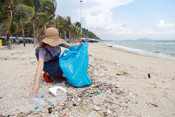 Volunteer tourist with hat is cleaning up garbage and plastic debris on dirty beach by collecting them into big blue bag