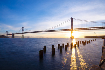 Fototapeta na wymiar San Francisco Bay Bridge with light beams illuminating the blue waters at sunrise