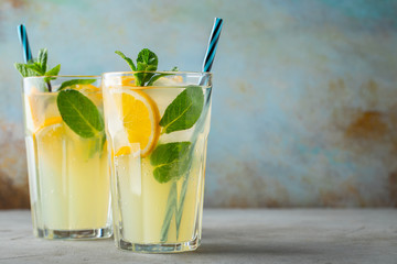 Two glass with lemonade or mojito cocktail with lemon and mint, cold refreshing drink or beverage with ice on rustic blue background. Copy space