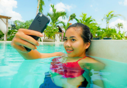 young happy and beautiful Asian Indonesian teen girl in bikini taking selfie picture with mobile phone in beach hotel swimming pool relaxed at tropical resort