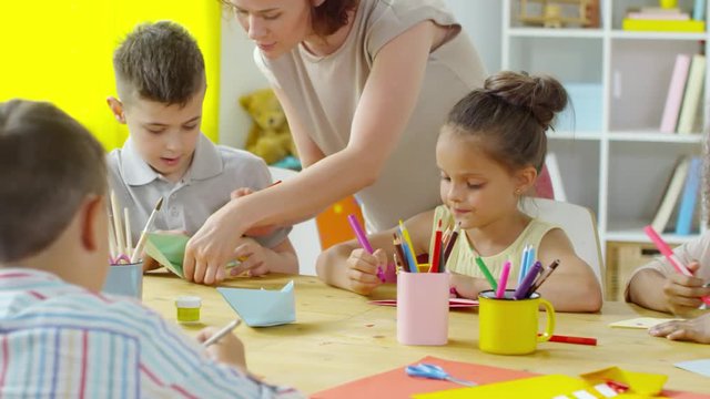 Panning shot of multi-ethnic little kids drawing on handmade origami boats with help of female teacher in art class