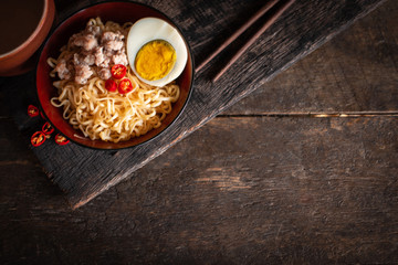 Instant noodle with pork, egg and vegetables on black bowl on the wood table there are chilli, chopstick and drinking glass placed around.