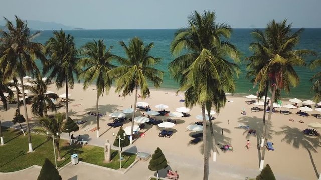 Aerial forward between coconut trees paradise beach white sand city promenade coastal exotic resort blue sea ocean waves tropical seascape.