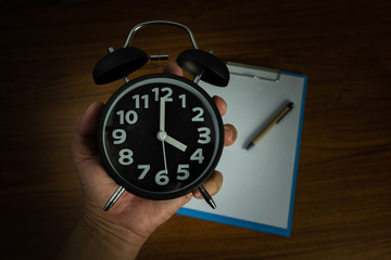 Naklejka premium Black vintage alarm clock in hand with clipboard and blank white paper and pen, copy space for add your text, work on time or over time and deadline concept.
