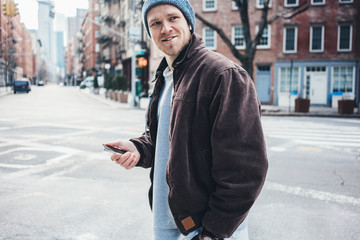 Young smiling man wearing hat and jacket walking on the city street with mobile phone in hand