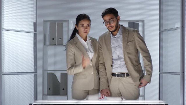 Asian businesswoman walking in office with middle eastern male colleague, turning on virtual multi-touch screen and showing presentation on it. Video suitable for adding AR graphics