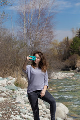 Young beautiful girl on nature takes a selfie on the phone and takes pictures of nature. Against the background of the forest and a beautiful mountain river. Tourism and travel.