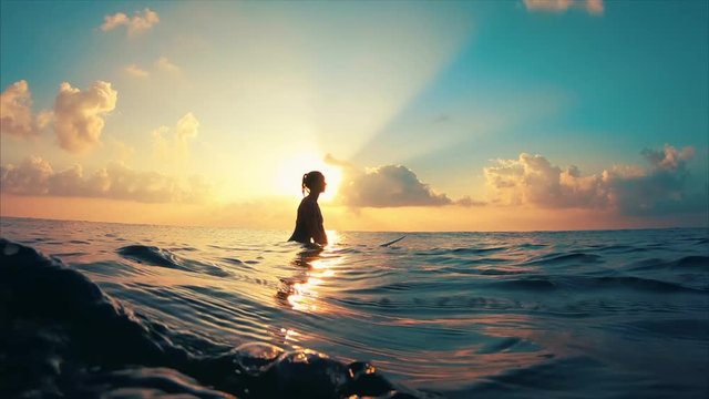 Young causasian woman surfer sits on the line up during morning surf session, waits her wave and enjoys warm tropical sunrise. Woman also looks at the camera and smiles. Clip contains underwater views