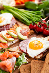 Huge healthy breakfast with sun-dried tomatoes, sandwiches with scrambled eggs, sausage, salmon, arugula, curd cheese, avocado on a wooden board