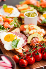 Huge healthy breakfast with sun-dried tomatoes, sandwiches with scrambled eggs, sausage, salmon, arugula, curd cheese, avocado on a wooden board