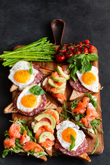 Mediterranean snacks set with brushettas, cheese variety, salami, eggs and salmon over black background on wooden board, top view