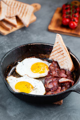 Fried eggs in a frying pan with sun dried cherry tomatoes, bacon and toasted bread for breakfast on a rustic wooden background.